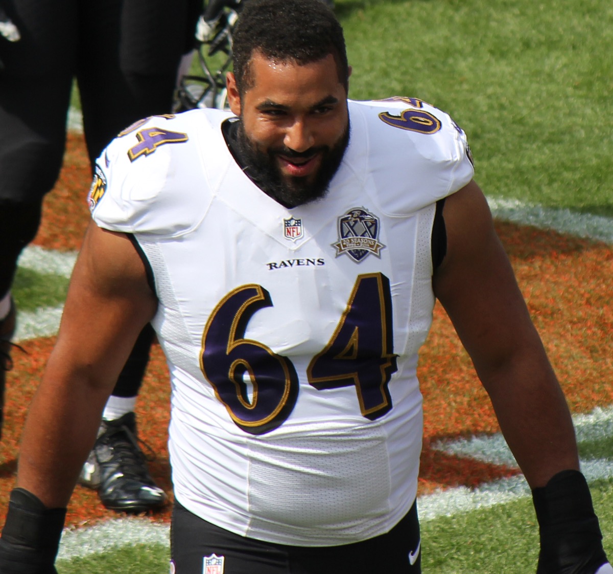 large football player in ravens jersey with number 64 smiling and walking toward camera on black background