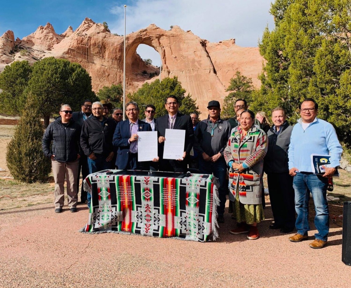 a group of people from the navajo nation standing outside in front of a beautiful red, orange natural sandstone archway. two men in the center hold up signed papers. they are standing behind a table