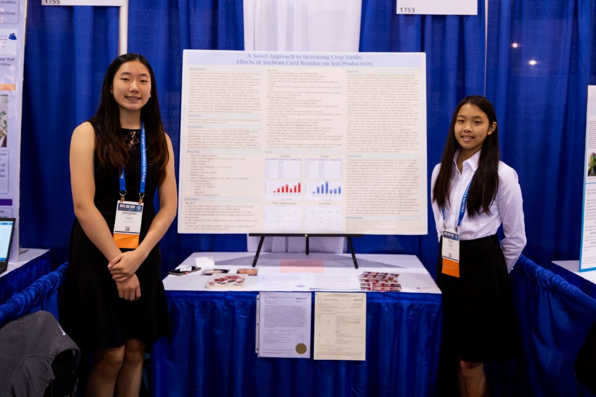 two asian students in front of a science board project. the student on the left is wearing a black dress and the student on the right is wearing a white shirt and skirt