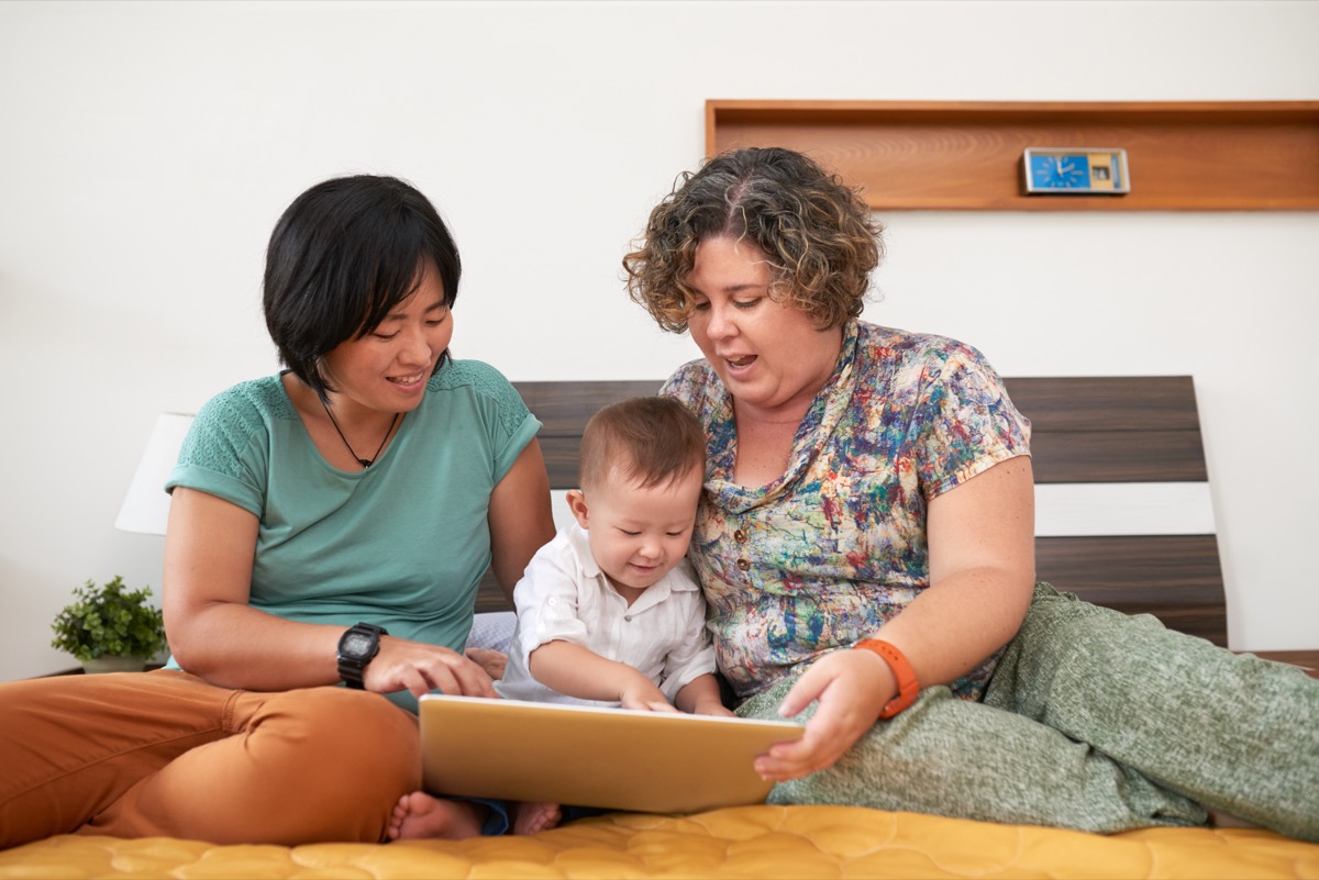 two women, one east asian, one white, sitting on a bed with their year-old child. all three are looking at a tablet computer.
