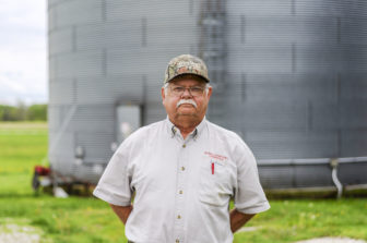 a farmer stands in front of a large silo looking into the camera