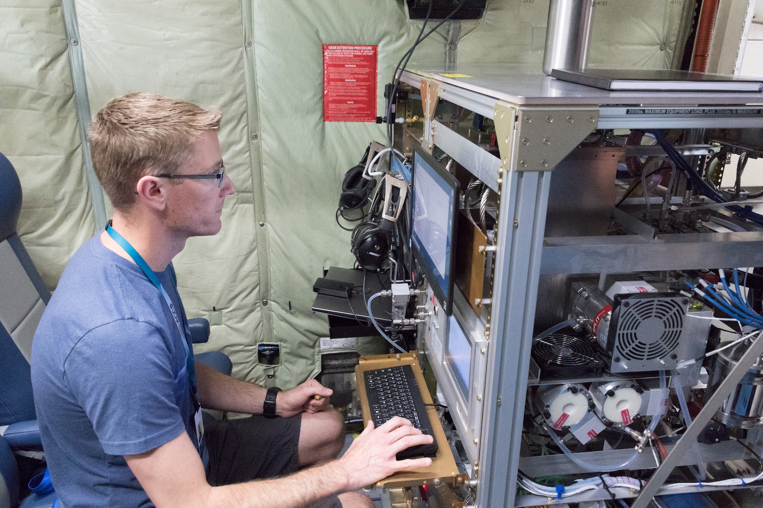 a man types at a large work station with exposed electrical system onboard a plane