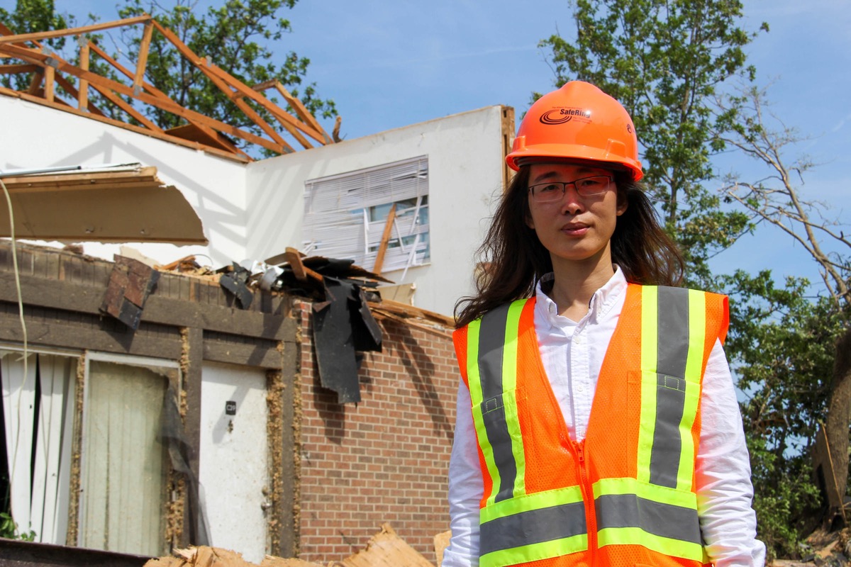 a woman in a hard hat stands in front of a tornado-damaged house