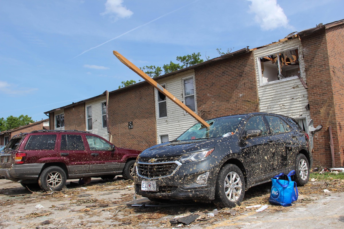 Cars in front of a damaged apartment building. both cars are covered in mud. a 2 by 4 wood beam has gone through the windshield of one car.