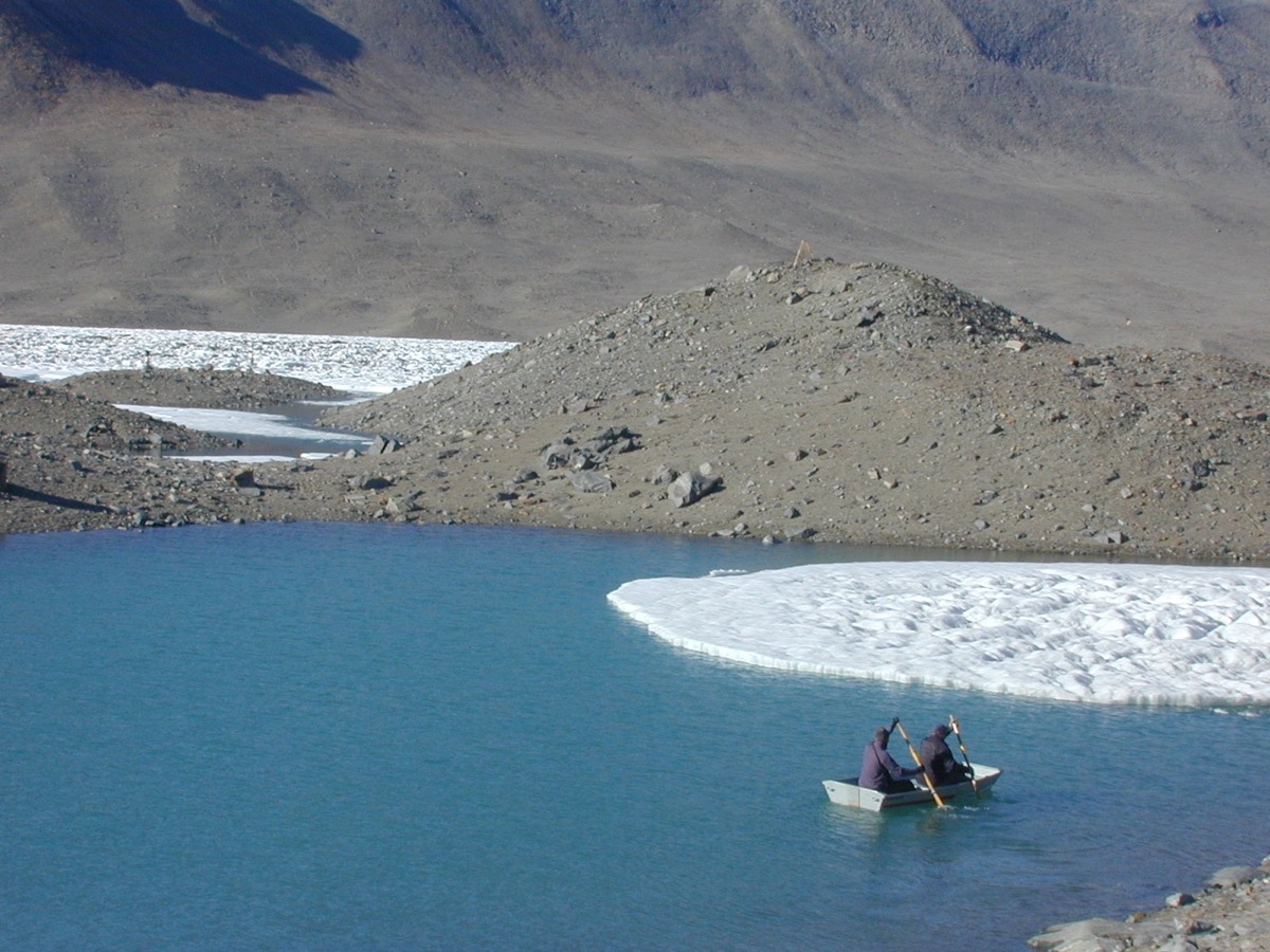 a large blue lake with some bits of snow near by but mostly barren rock. two people sit in a row boat