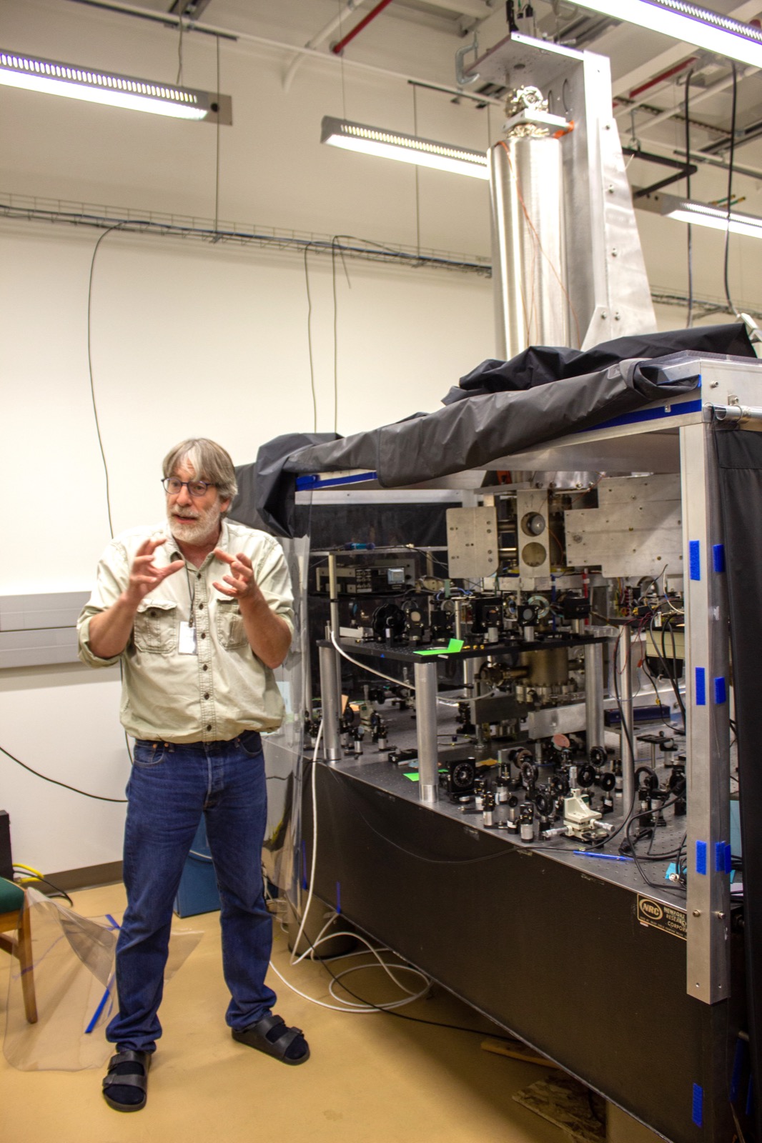 a man standing in front of large mechanical device with a pipe connecting it to the ceiling. a side panel is open, revealing its electronic guts