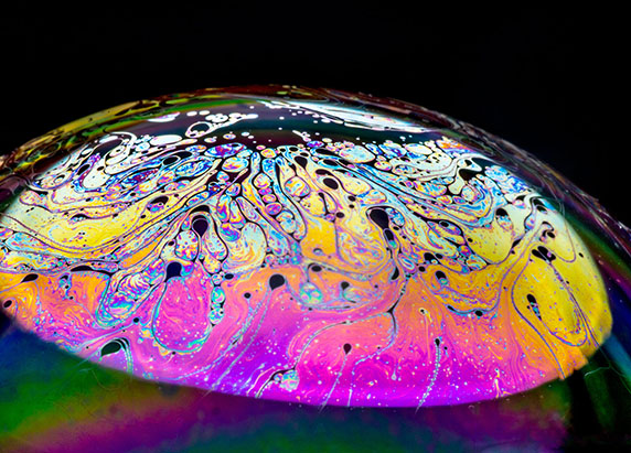 a close up of a surface of a bubble. the surface is smooth and concave and is swirled with bright shiny magenta, yellow, blue, purples, and green.
