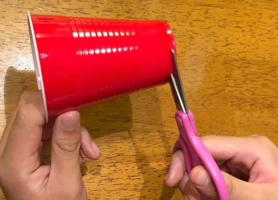 a hand holds a red solo cup on its side while the person cuts the bottom of the cup with scissors