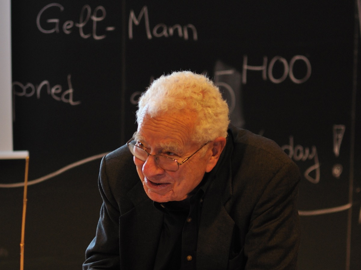 murray gell-mann, an older man with white hair and glasses, standing in front of a blackboard with equations
