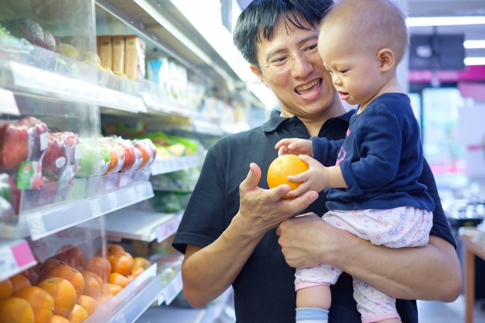 a smiling dad hands his infant an orange in a grocery store