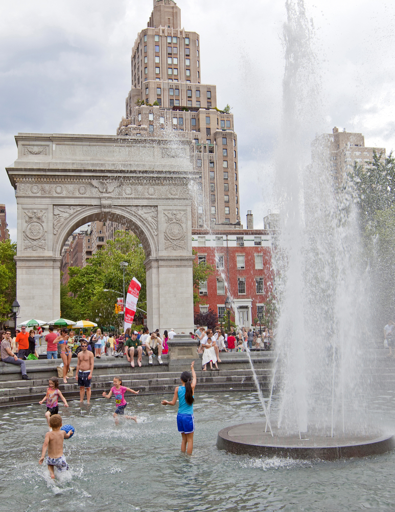 four kids run and play in a large fountain. there is a french architectural arch in the background