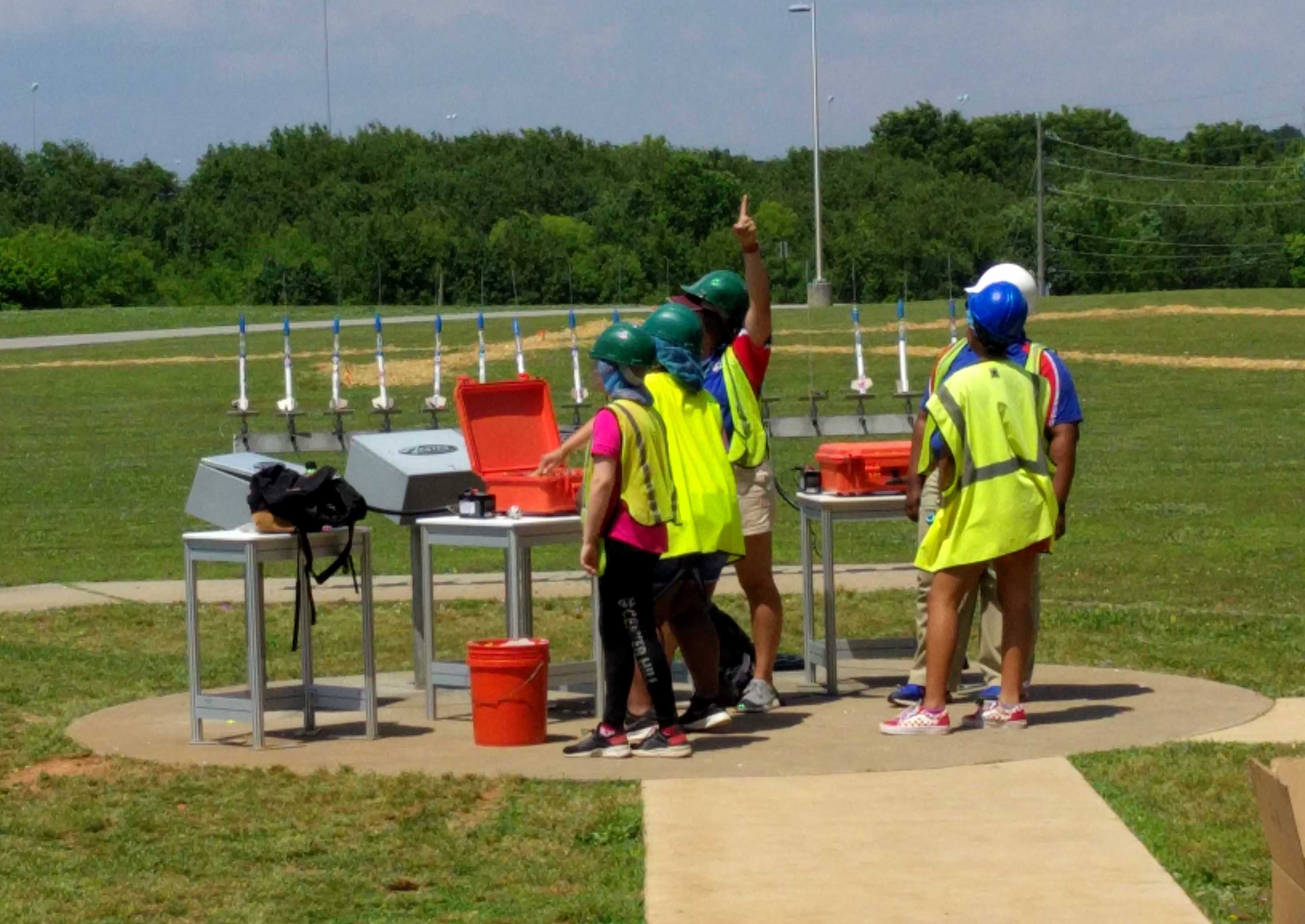 five kids in hard hats and safety vests stand in a grassy field with a row of model rockets