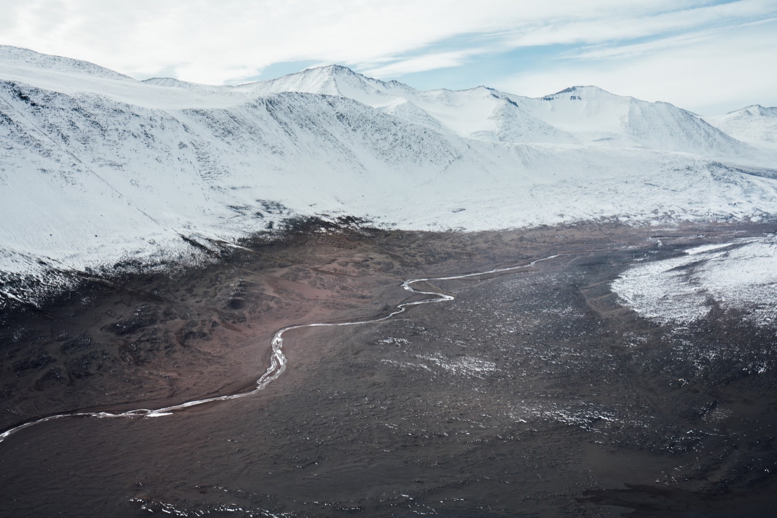 an ariel shot of a winding stream in a valley between snowcapped mountains