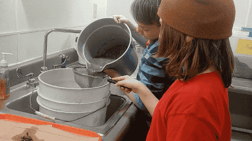 a kid in a red shirt watches a kid in a blue striped shirt pour a muddy liquid through a sieve over a sink