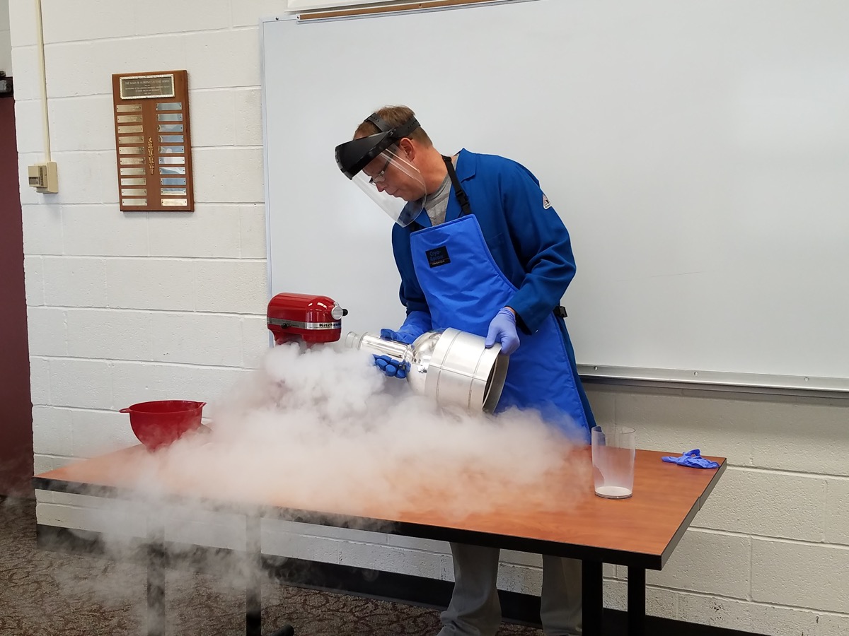 a man with a protective face visor, apron, and gloves, pours liquid nitrogen in a kitchen mixer. fog spills over the table