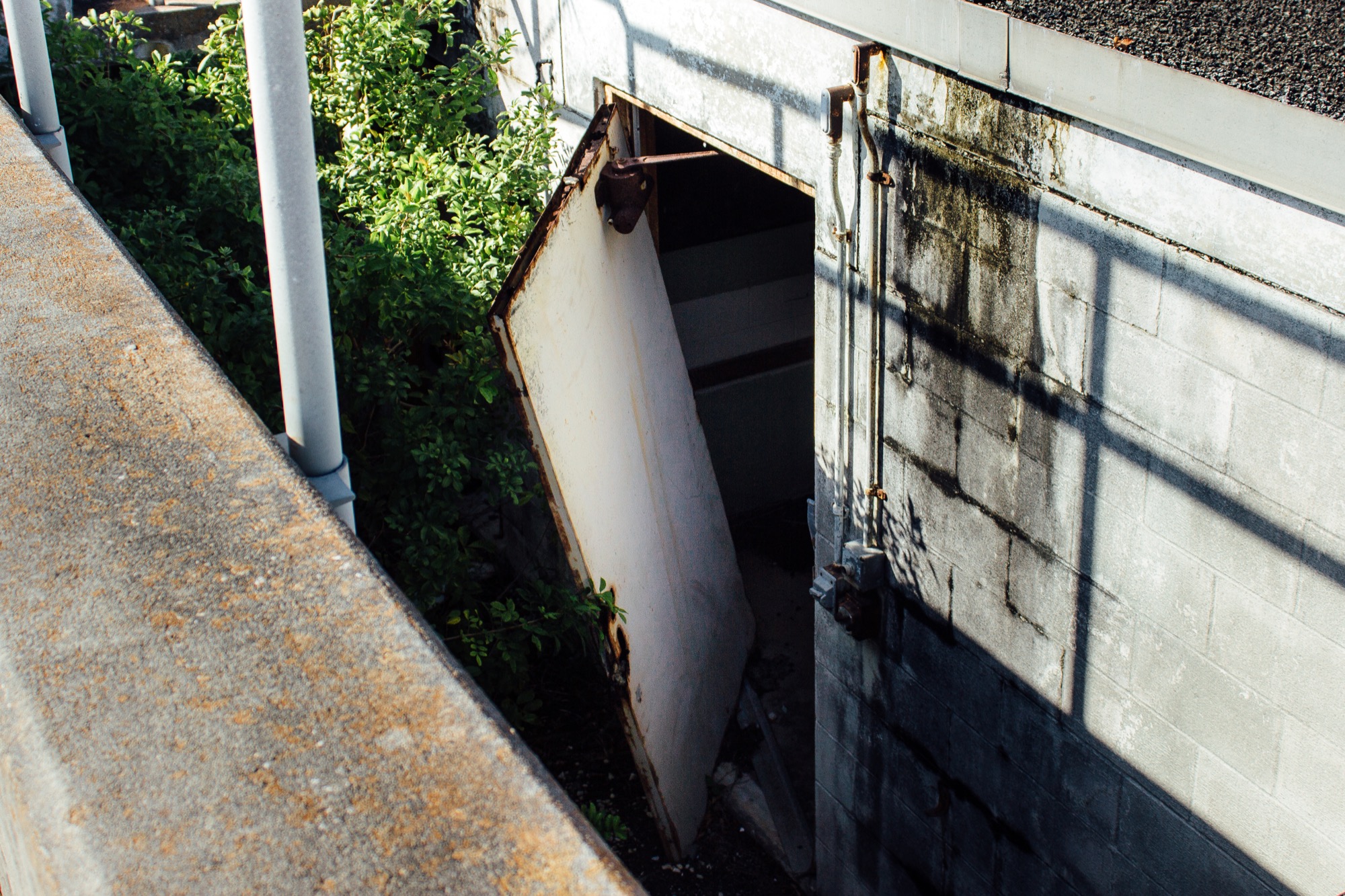 a rusted, broken door is ajar, hanging by only one of its hinges, attached to a squat rusted building