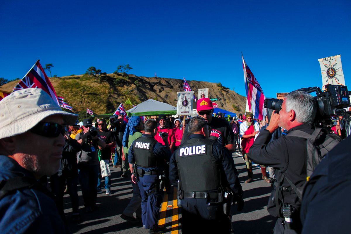 a group of protesters/protectors hold signs and hawaiian flags. there are police walking and patrolling the crowds