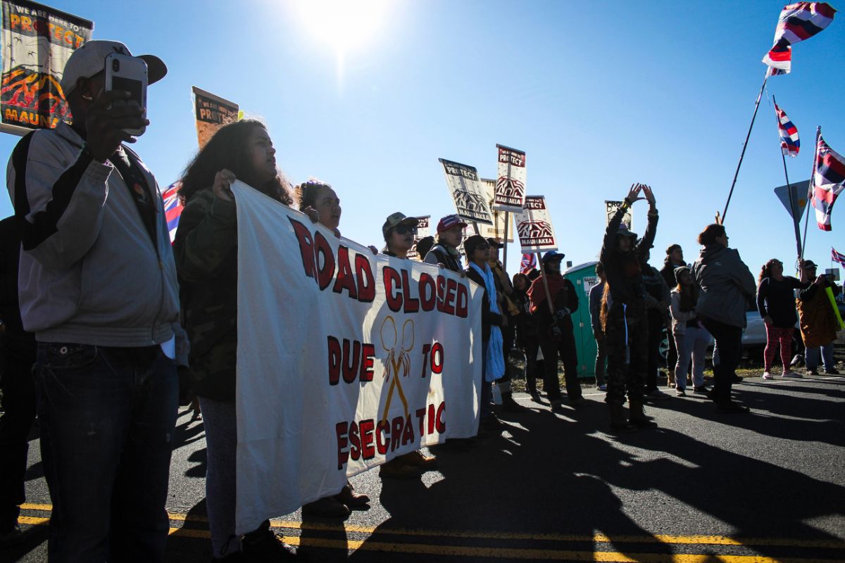 a large group of protesters/protectors hold flags and signs on mauna kea. one large sign reads 'road closed due to esecration'