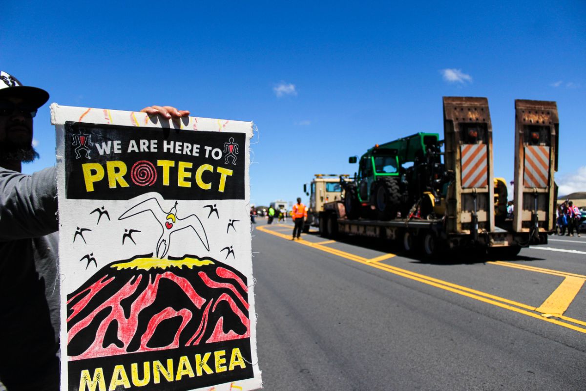 a protester/protector holds a sign that reads 'we are here to protect mauna kea.' in the background is a large tractor and construction truck