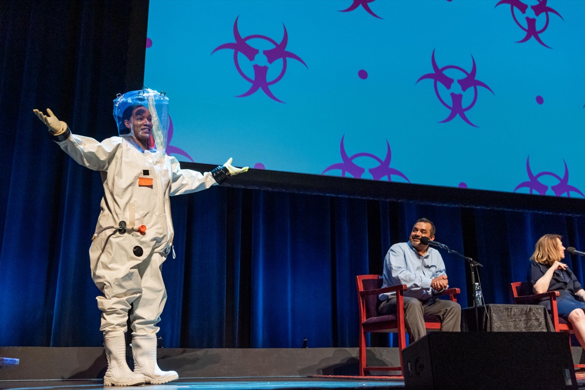 a woman stands on stage in an astronaut like suit. next to her sit two scientists with mics, talking to ira flatow