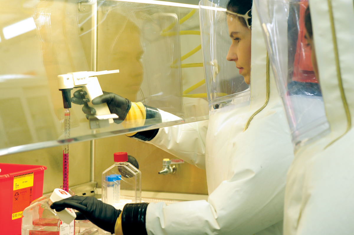 A scientist in a puffy safety suit. She is pipetting liquid into a vial.