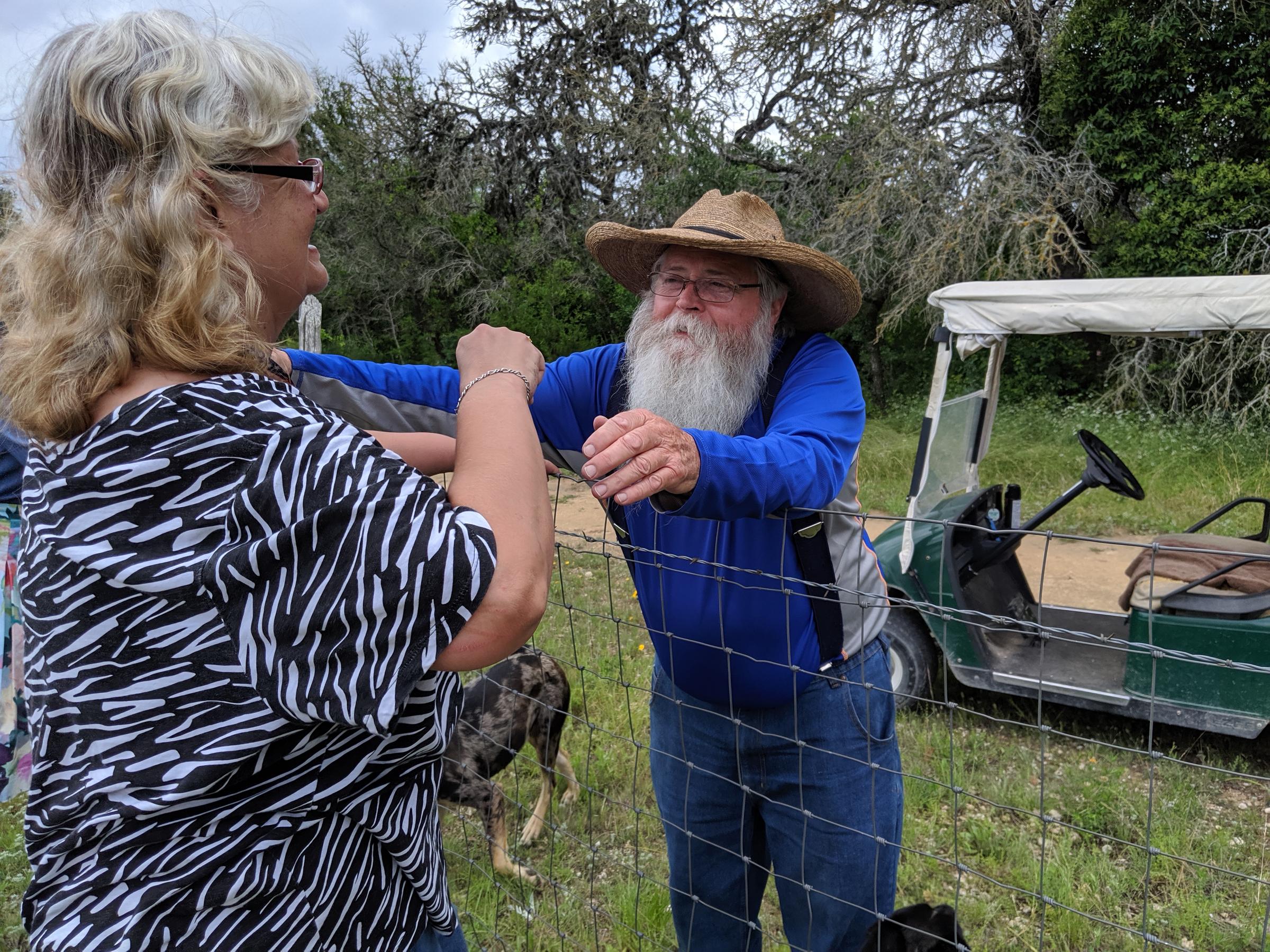 man with big white beard and giant hat leans to hug a woman over a wire fence