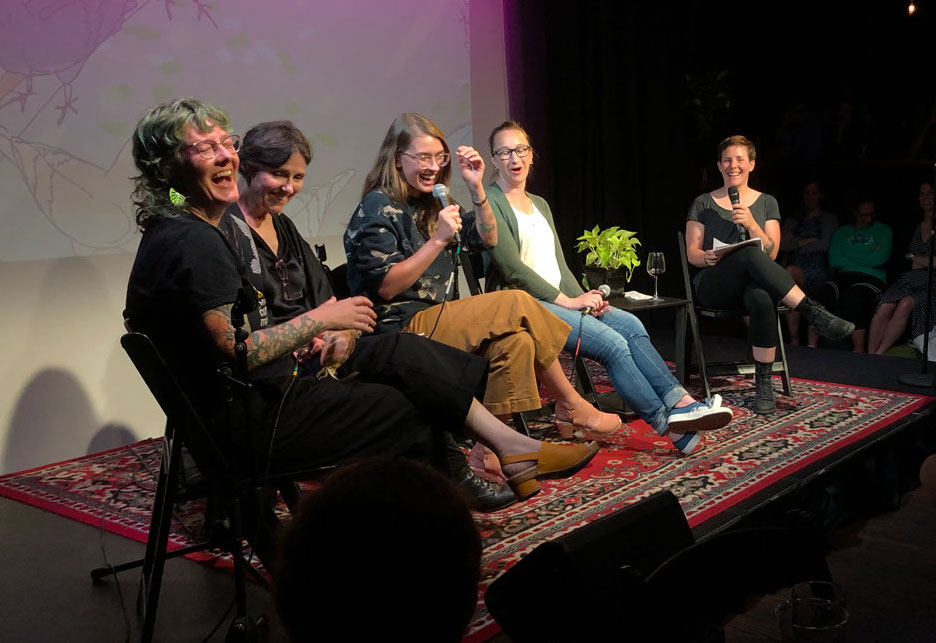 five women stage all with mics laughing with an illustrated bird background behind them