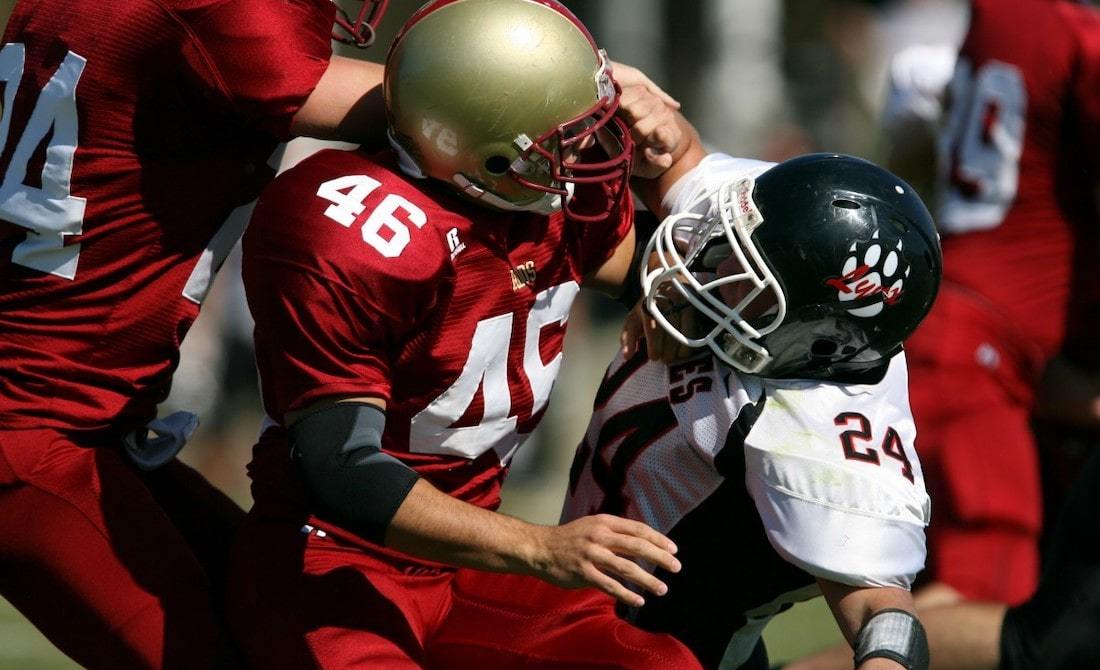 two football players - one in red and one in white, both wearing helmets - colliding and falling to the ground in the middle of a game, surrounded by other players