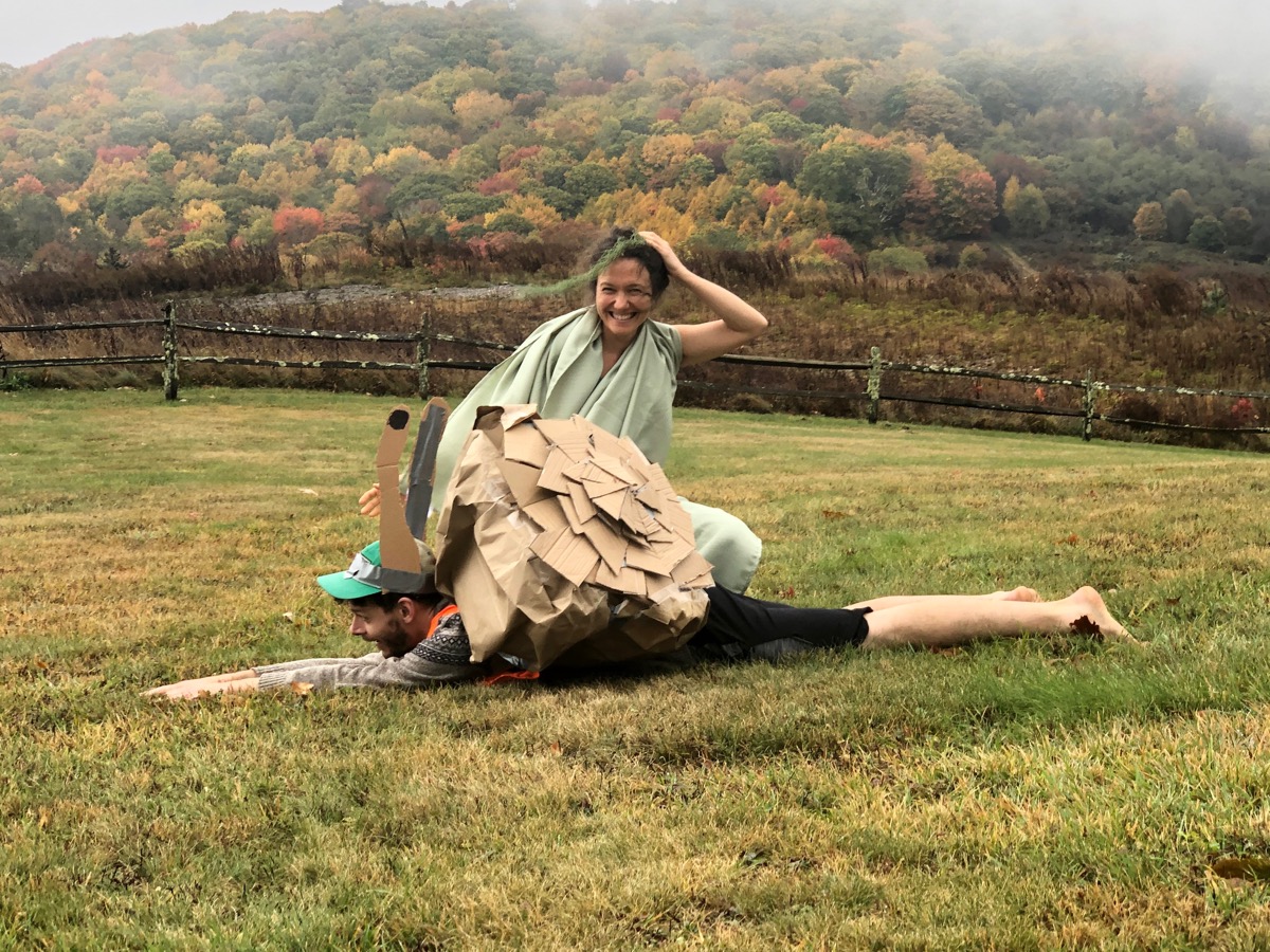 a man in a snail costume which is made of cardboard lies on the grass mimicking a snail, while a woman in a green table cloth dressed as a lichen stands behind him smiling