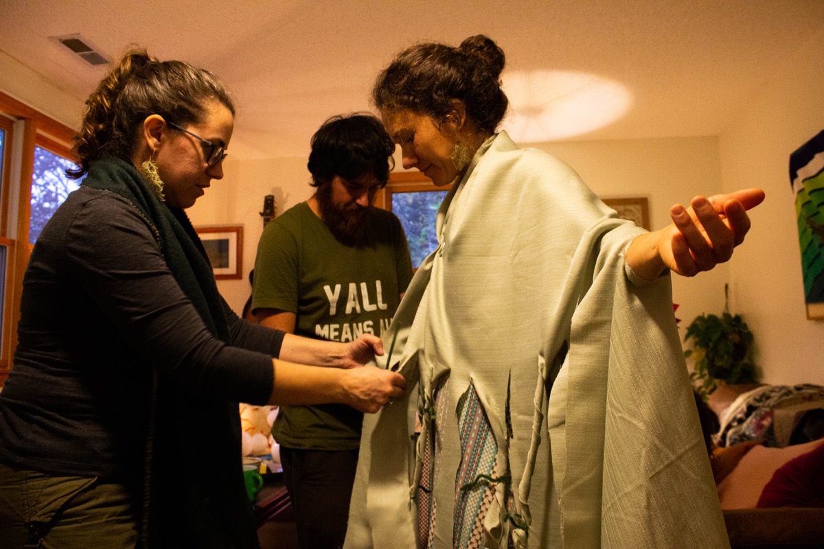 two people assist a woman creating a costume by tying strips of green table cloth