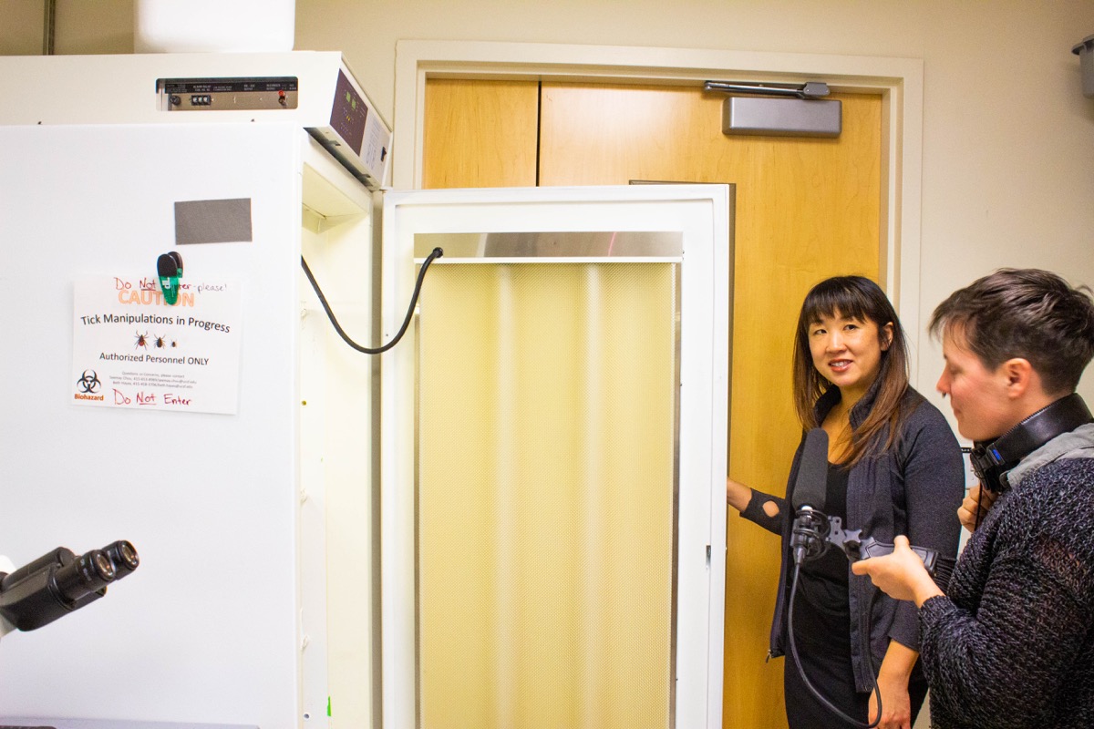 a large fridge with a sign that reads "caution tick manipulation in progress" while a woman opens the door, and another woman interviews her with a microphone