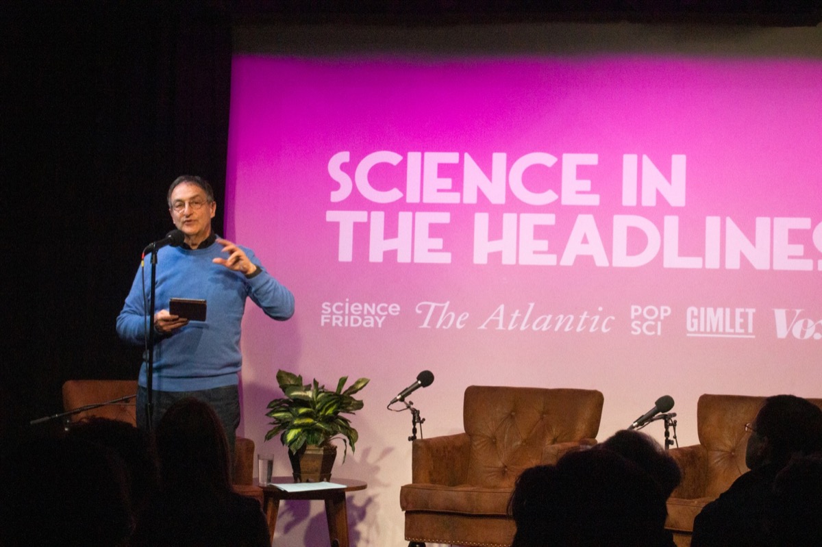 a man stands on stage. a slide behind him reads "science in the headlines"