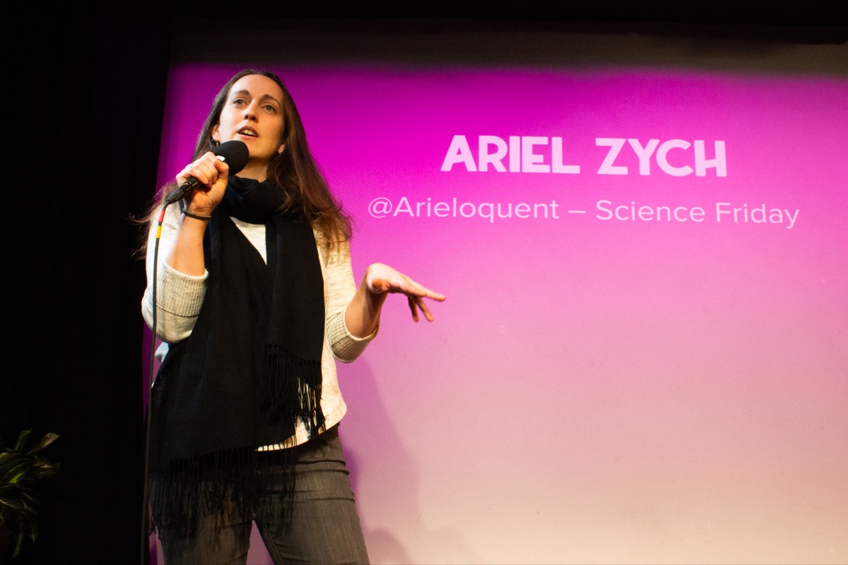 woman on stage with a microphone, with the name 'ariel zych' projected on a pink-tinted projection screen behind her