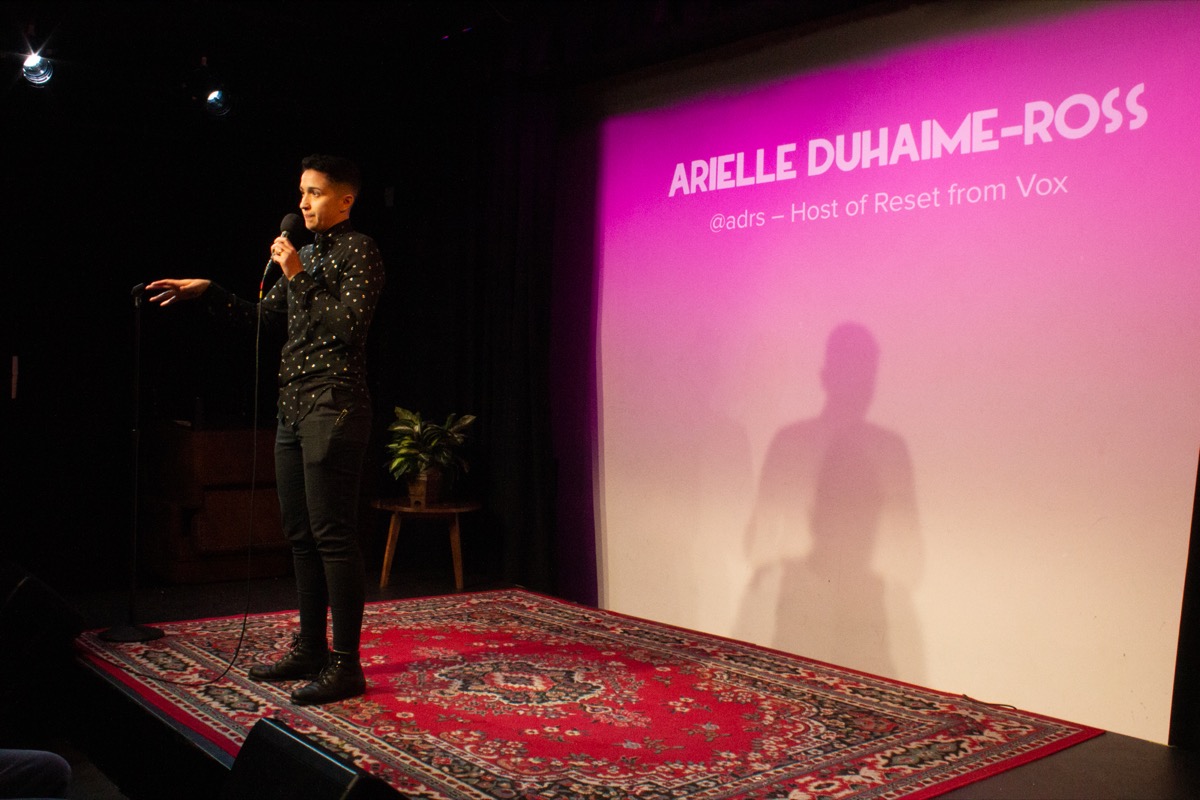 woman with a microphone on stage, with the name 'arielle duhaime-ross' projected on a pink-tinted projection screen behind her