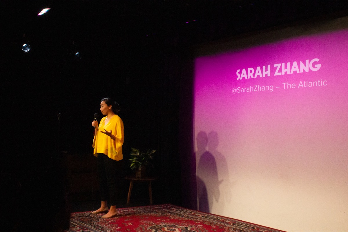 woman in a yellow shirt with a microphone on stage, with the name 'sarah zhang' projected on a pink-tinted projection screen behind her