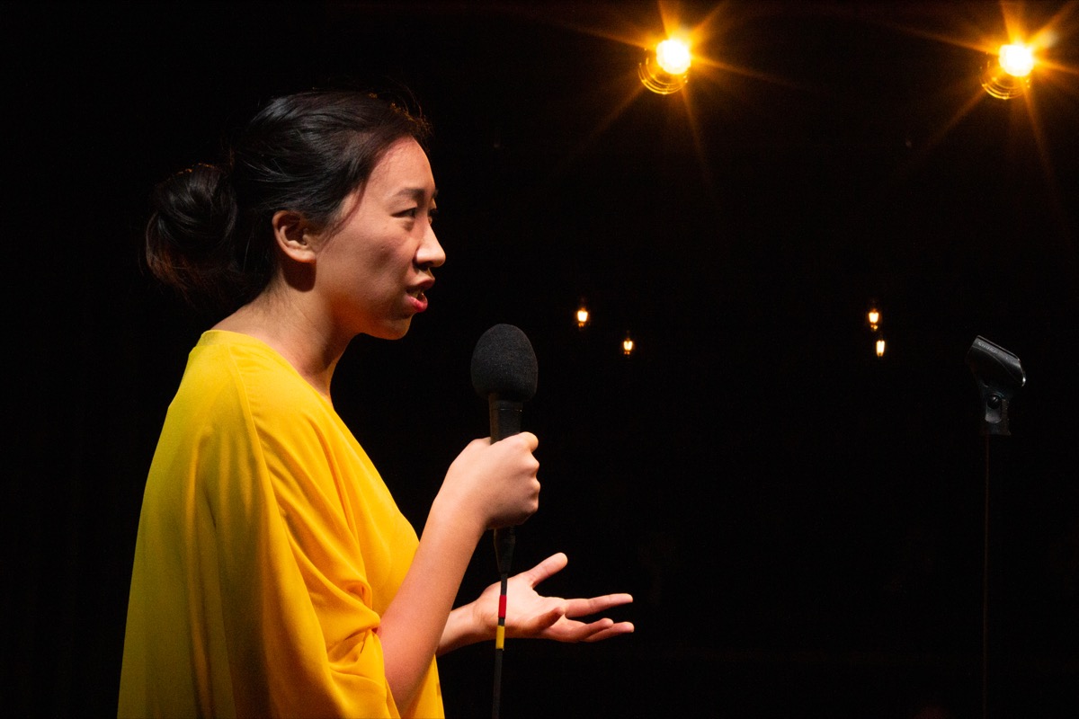 woman in a yellow shirt with a microphone, looking inquisitively and gesturing openly with her left hand toward the crowd