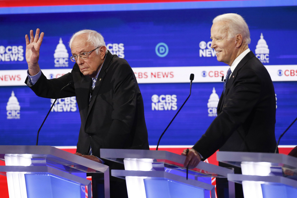 bernie sanders, raising his hand, on a debate stage with joe biden