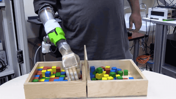 a person with a prosthetic hand picks up wooden blocks and drops them in another bin