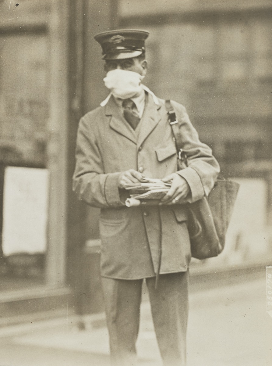 a black and white photo of a man in a flat cap and coat and a bag of mail. he wears a face mask