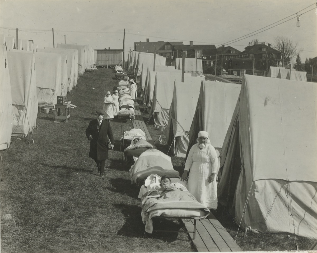 rows of white tents with beds and patients lying outside. nurses come by to check on them. in the background you can make out nearby homes