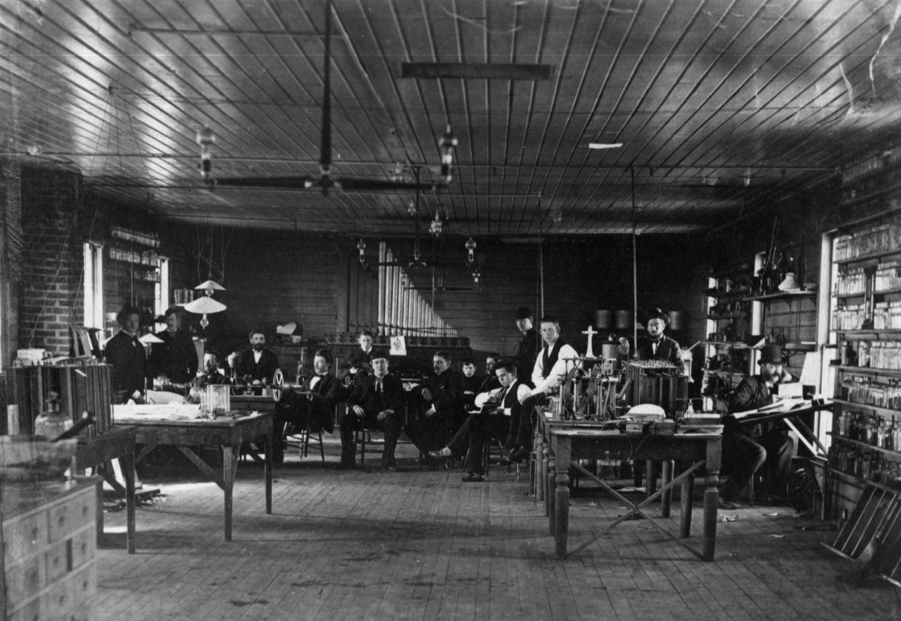 a black and white photo of a group of over a dozen people in a room with lightbulbs and chairs and tables