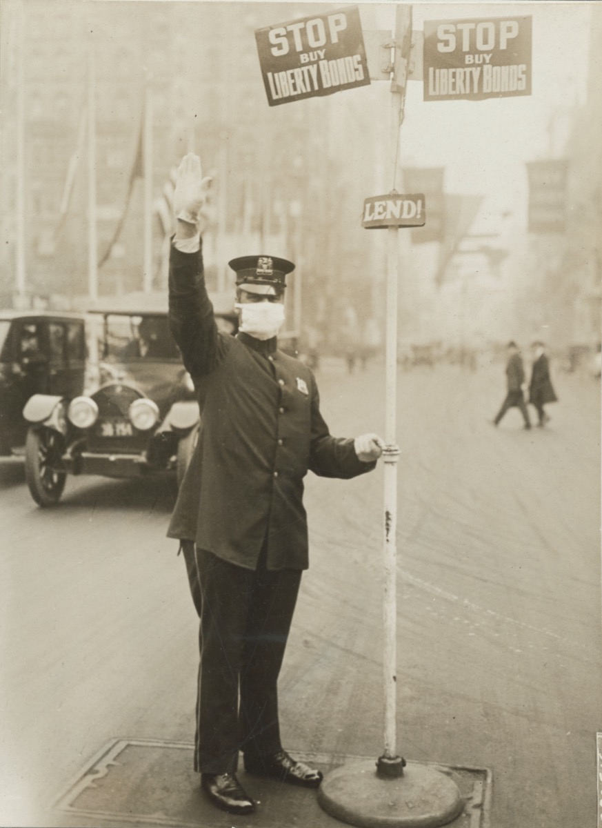 a man in a flat cap, white gloves, and face masks, directs car traffic