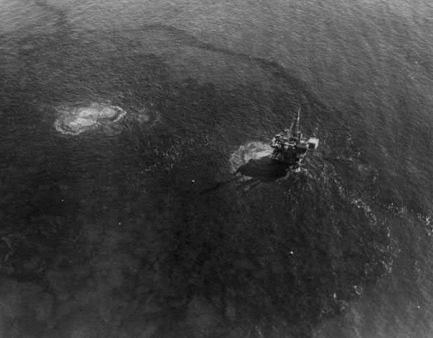 a black and white photo of an offshore oil rig seen from above, where you can see black oil bubbling and expanding out from the rig beneath the ocean