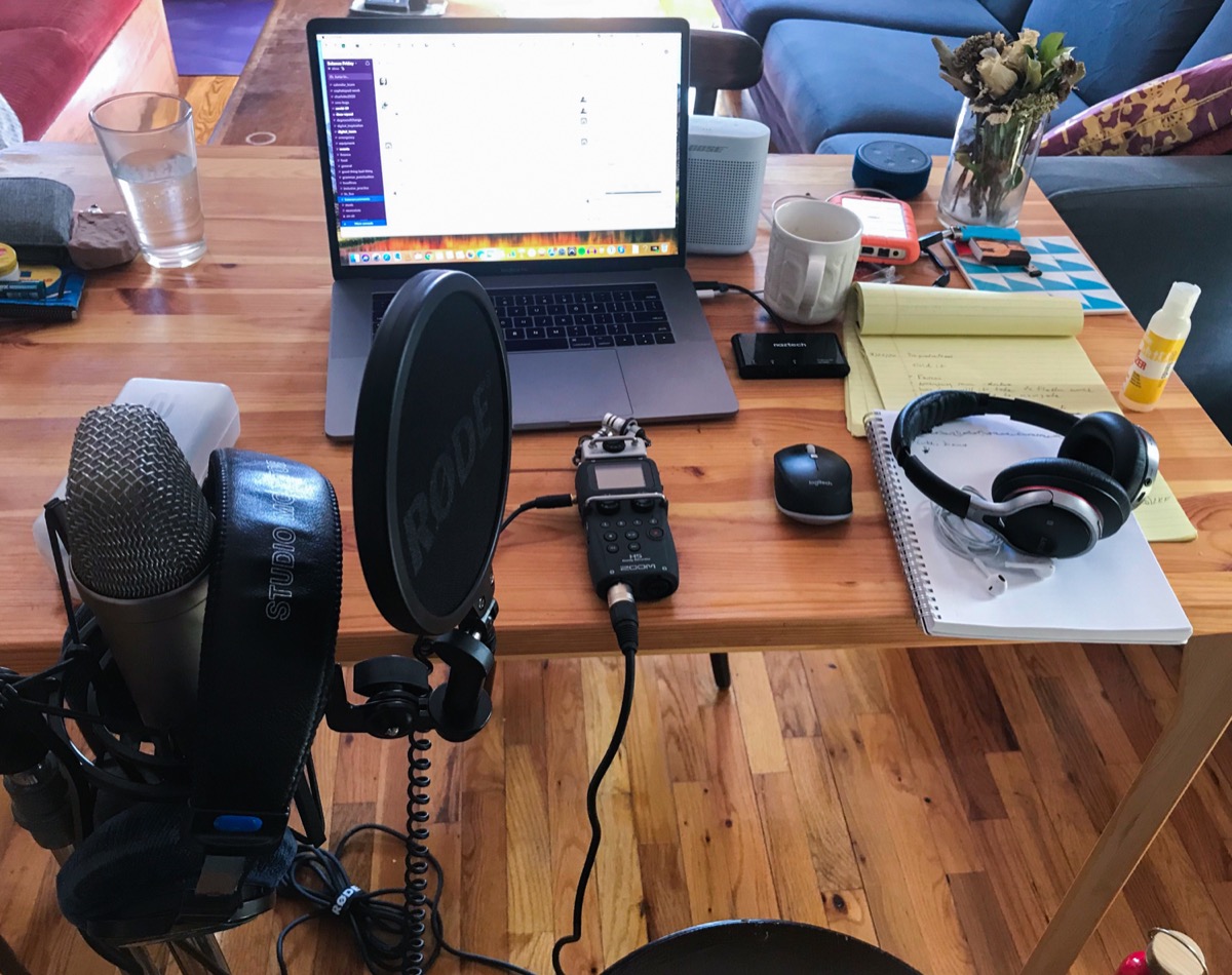 a wooden table at an apartment with a laptop, headphones, note pad, and an audio recorder. close up is a mic on a stand