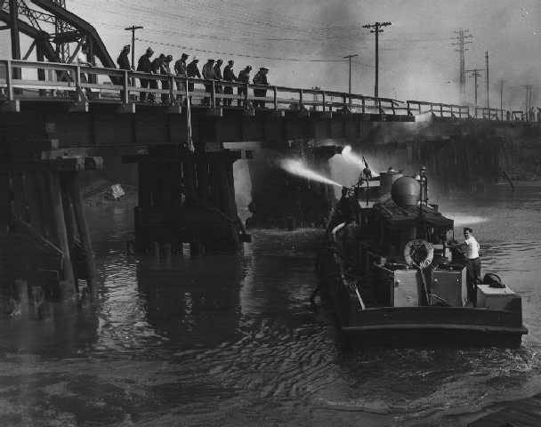 a boat on a river trying to put out a fire on the river. people watch on a bridge overhead