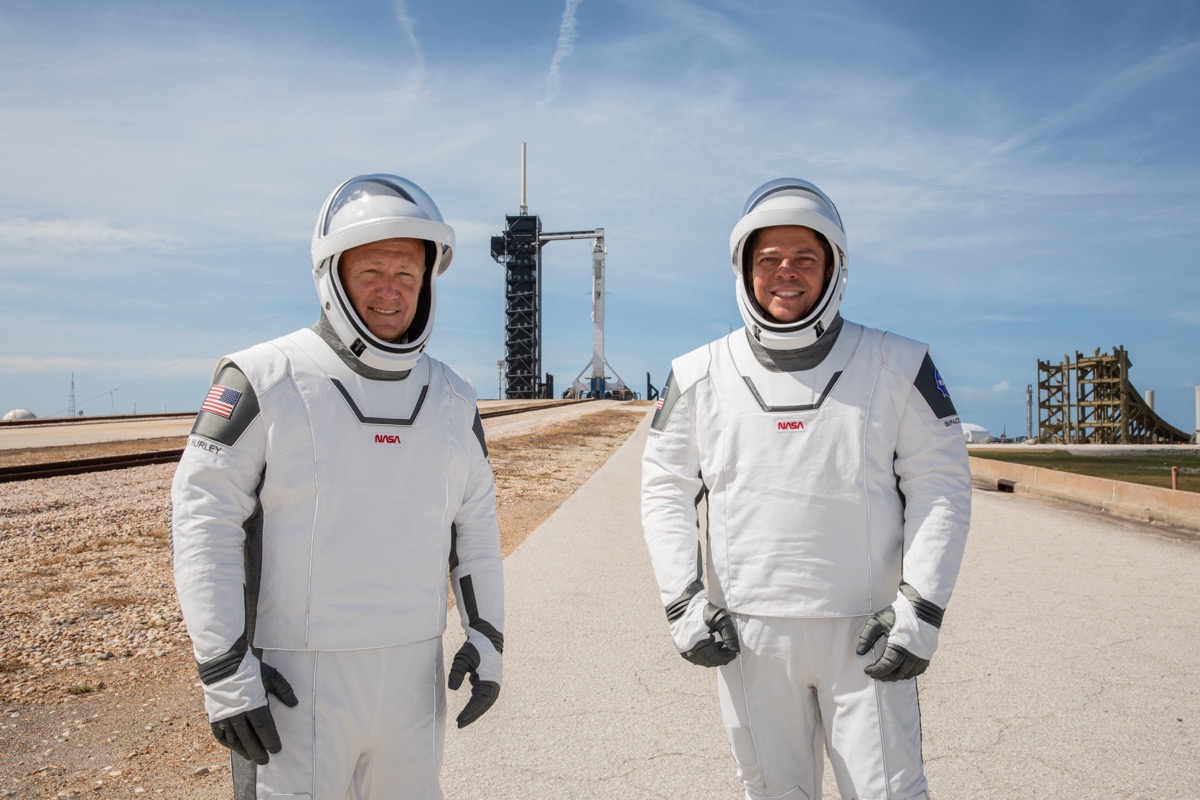 two men in space suit, flight gear on a landing pad. in the background is a rocket