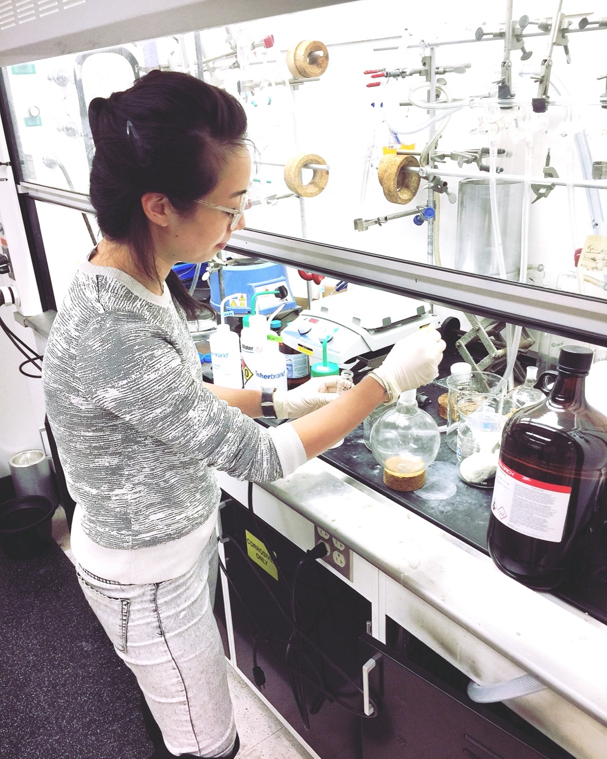 a woman wearing gloves in a chemistry lab in front of a lot of beakers, mixing a solution