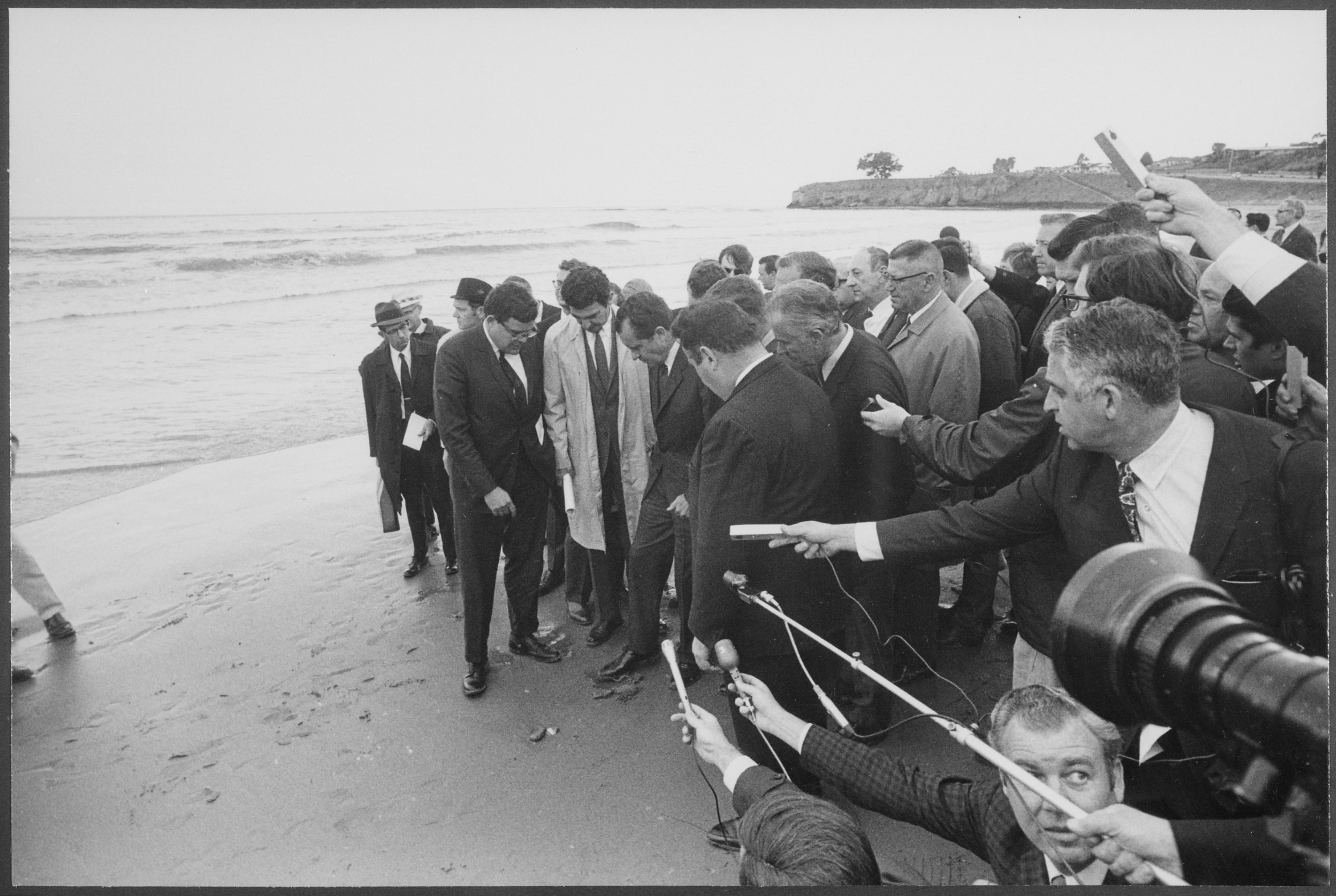 Группа репортеров следует за президентом Никсоном на пляже, упираясь ногами в залитый нефтью песок.