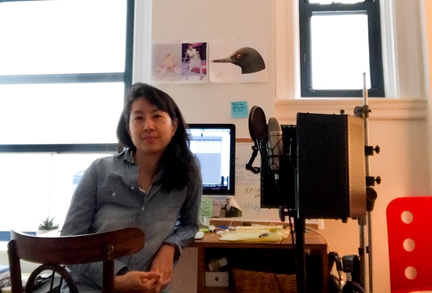 an asian woman sits at a desk at home with a mic stand and microphone next to her