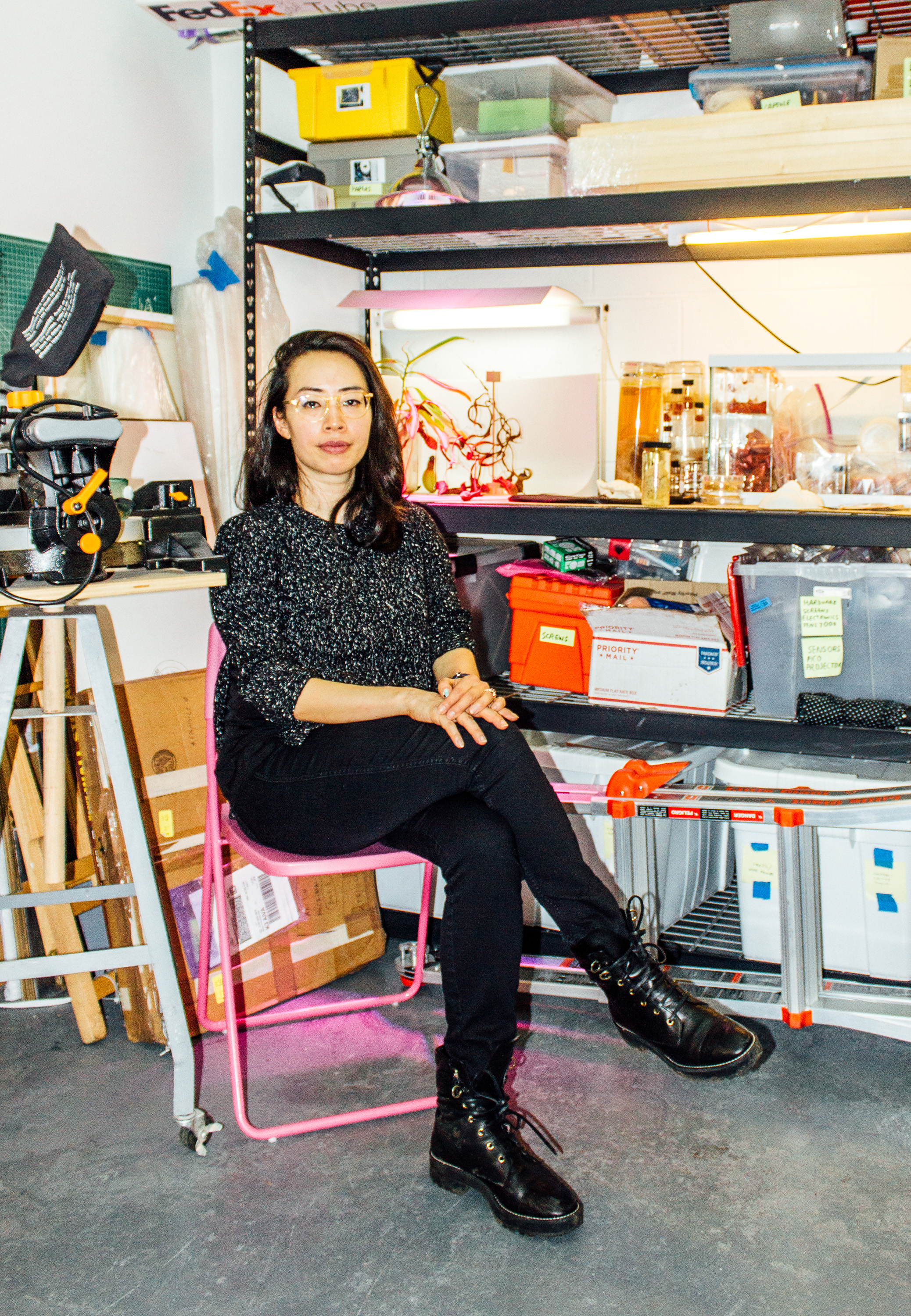 a woman sitting in a chair in a window-less studio, with shelves behind her containing plants under special light, and cylinders of body parts